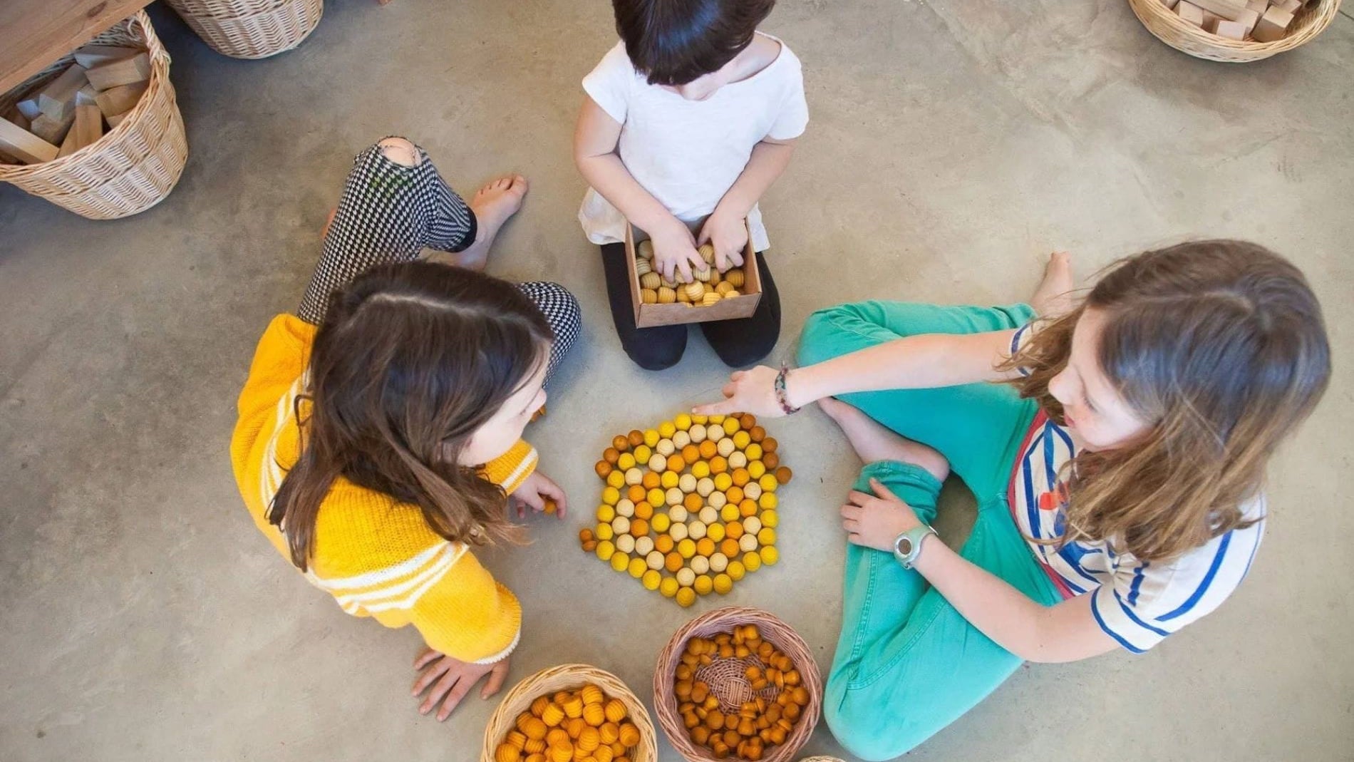 Three children gathered around a collection of baskets filled with oranges on a concrete floor.